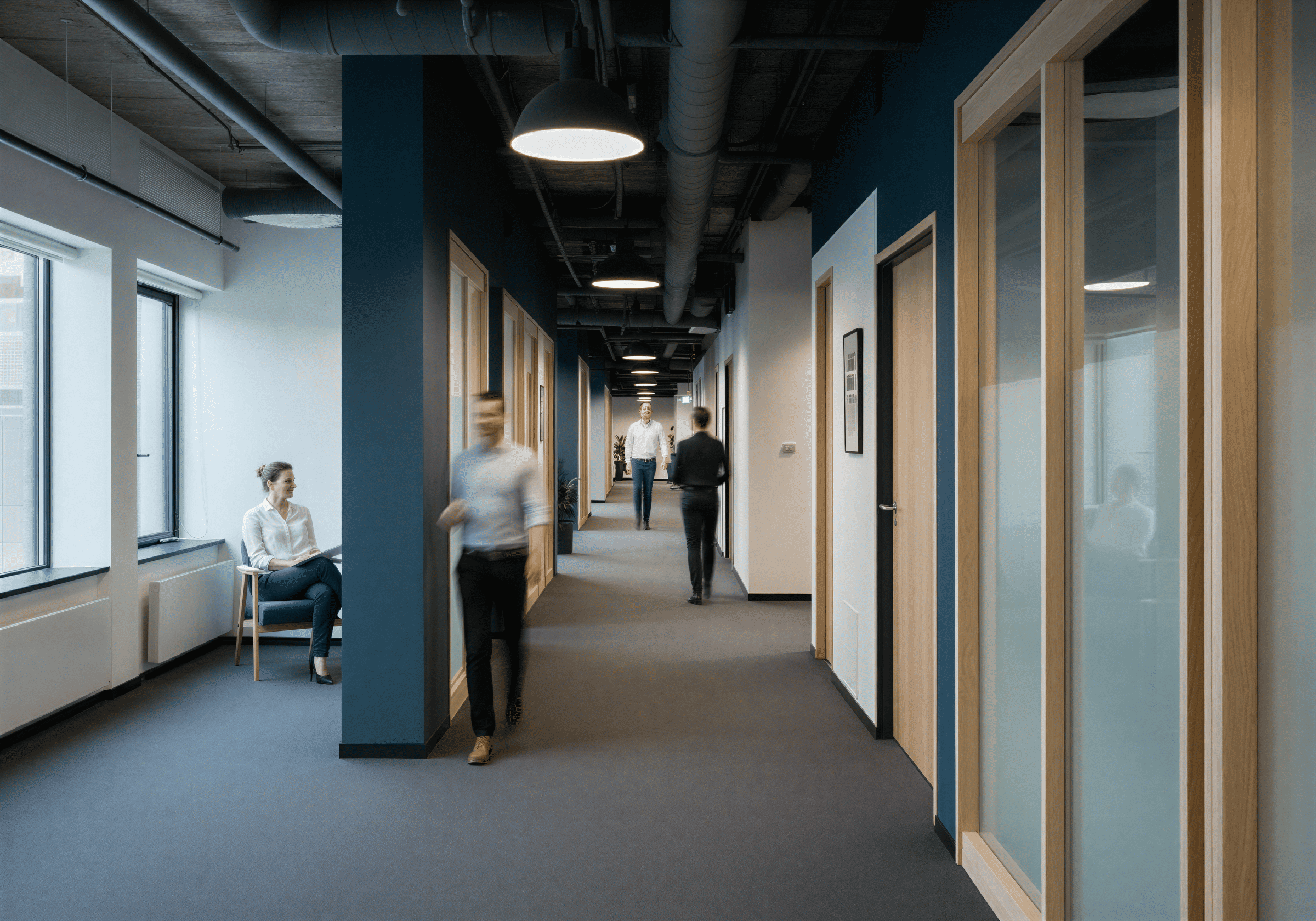 Modern office corridor with several people walking and a woman sitting by the window.