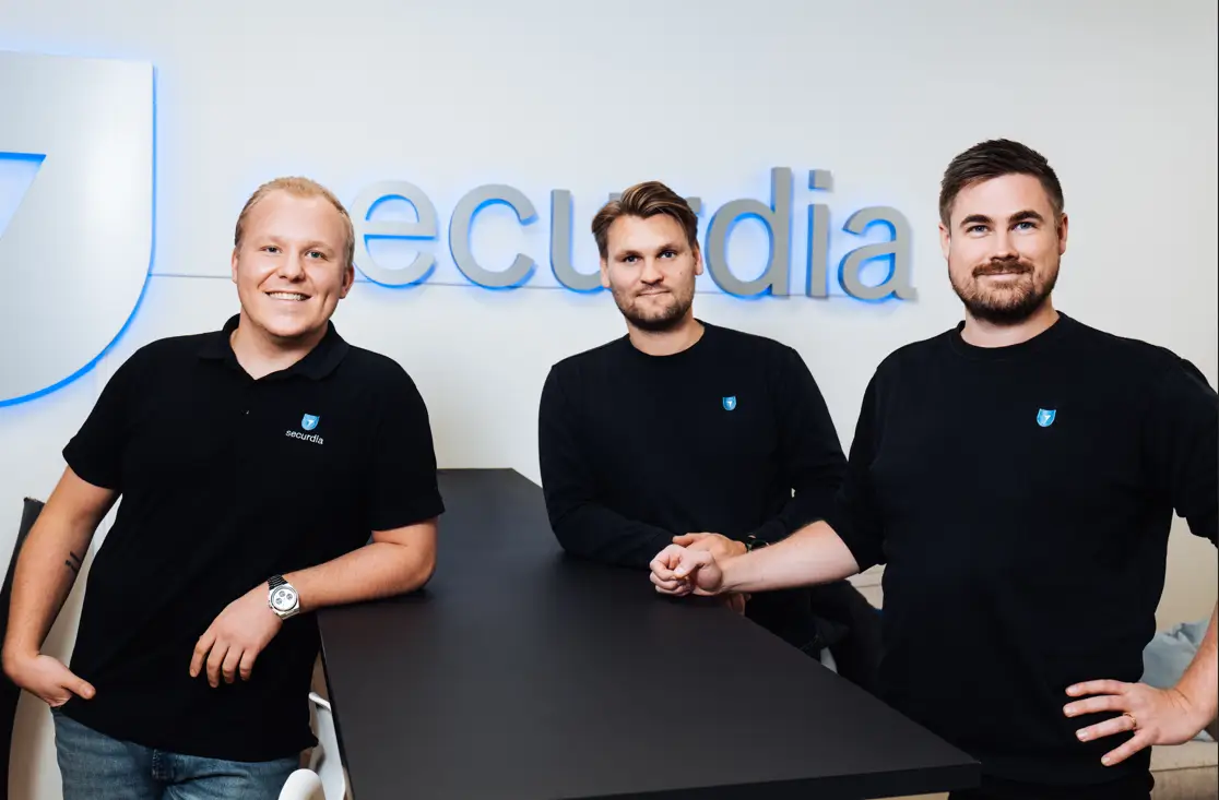 Three men pose in front of a wall with the text "securdia", all wearing black sweatshirts with the company logo.